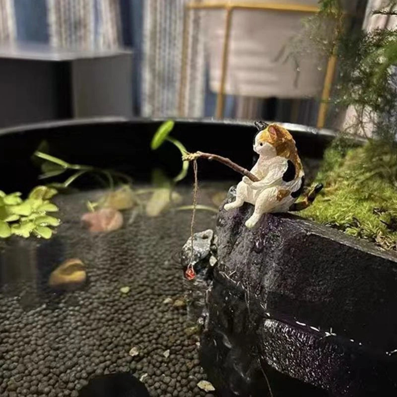 Decorative figurine of a cat fishing in a small pond with plants and rocks.