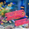Two red cylindrical fish tanks with a lattice cutout design, shown in an aquarium setting with a live betta fish swimming through the top one and a coral stand supporting the bottom one.